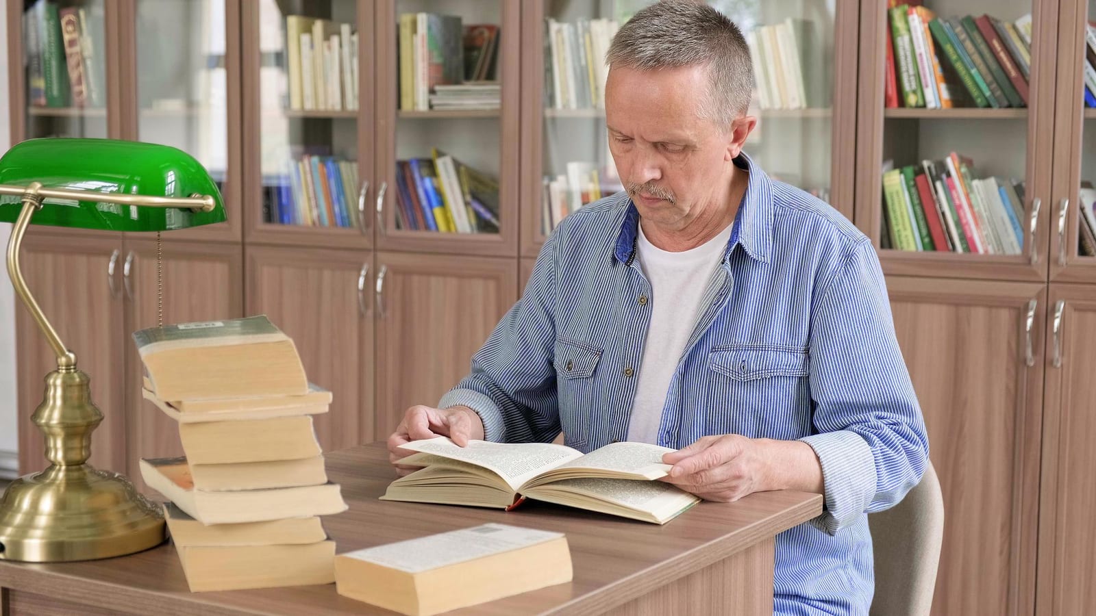 Homem lendo um livro aberto sob a luz de uma luminária verde de mesa, com uma pilha de livros ao lado e estantes cheias ao fundo.