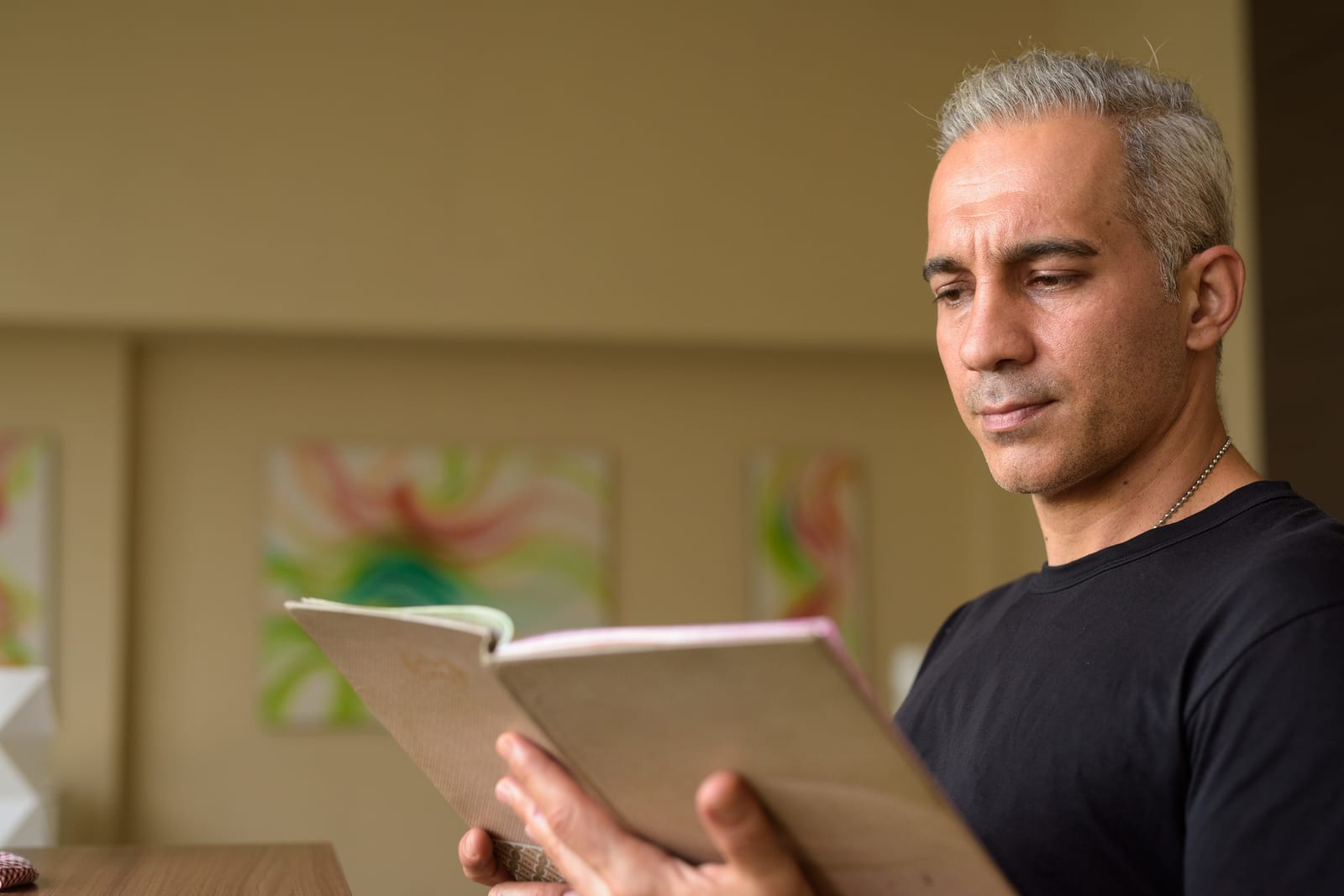 Homem segurando um livro aberto de capa neutra, lendo atentamente em ambiente iluminado.